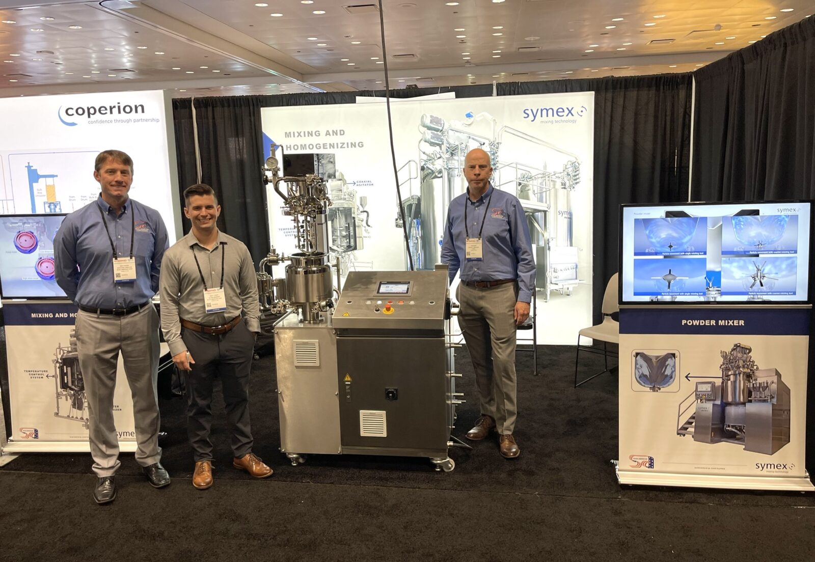 Ben Parker, Alec Tussey, and Tim Kruse standing in the symex booth at INTERPHEX 2022 with symex equipment, signage, and TV displays in the background.